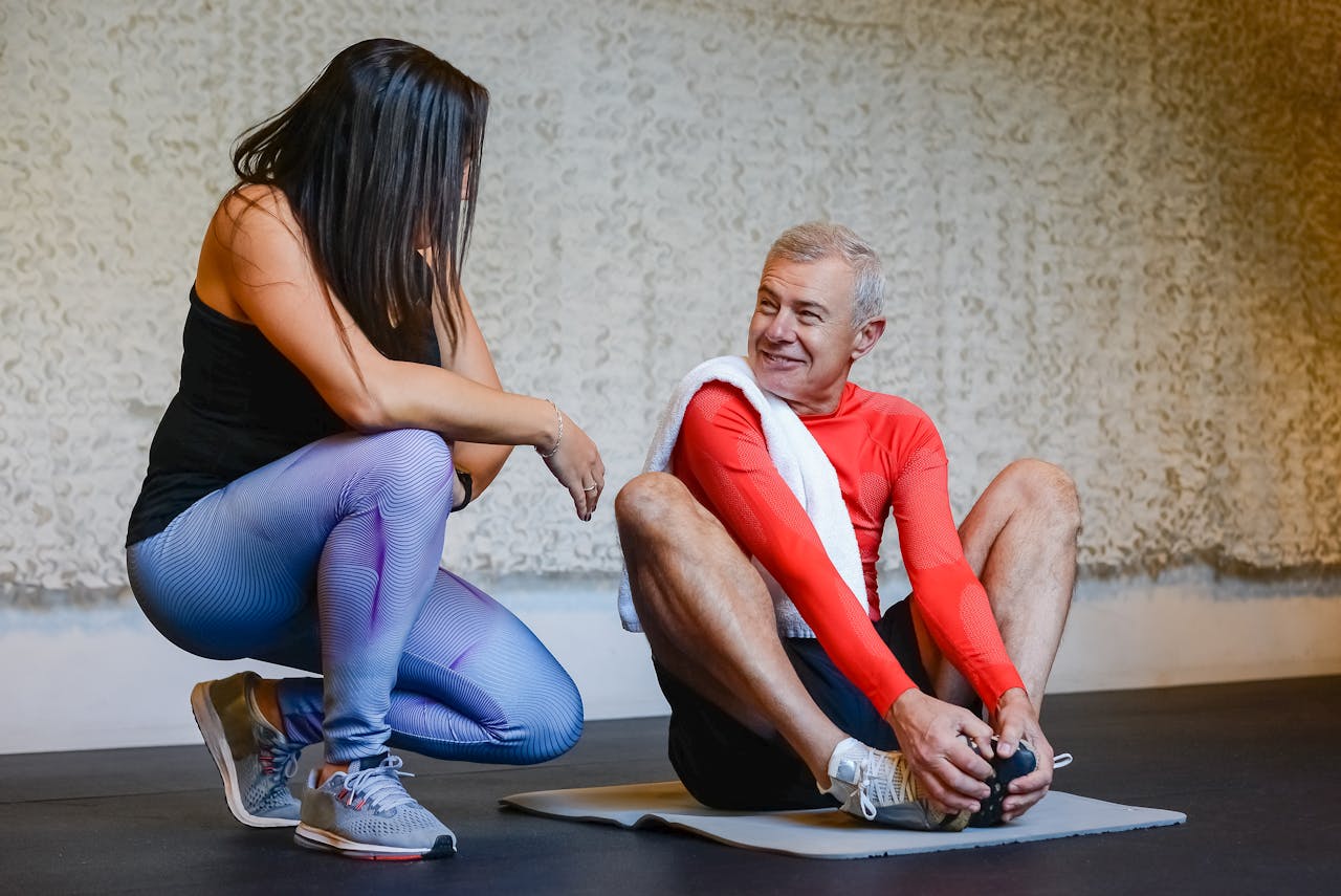 Fitness coach helps senior man stretch on exercise mat in a gym setting, promoting health and wellness.
