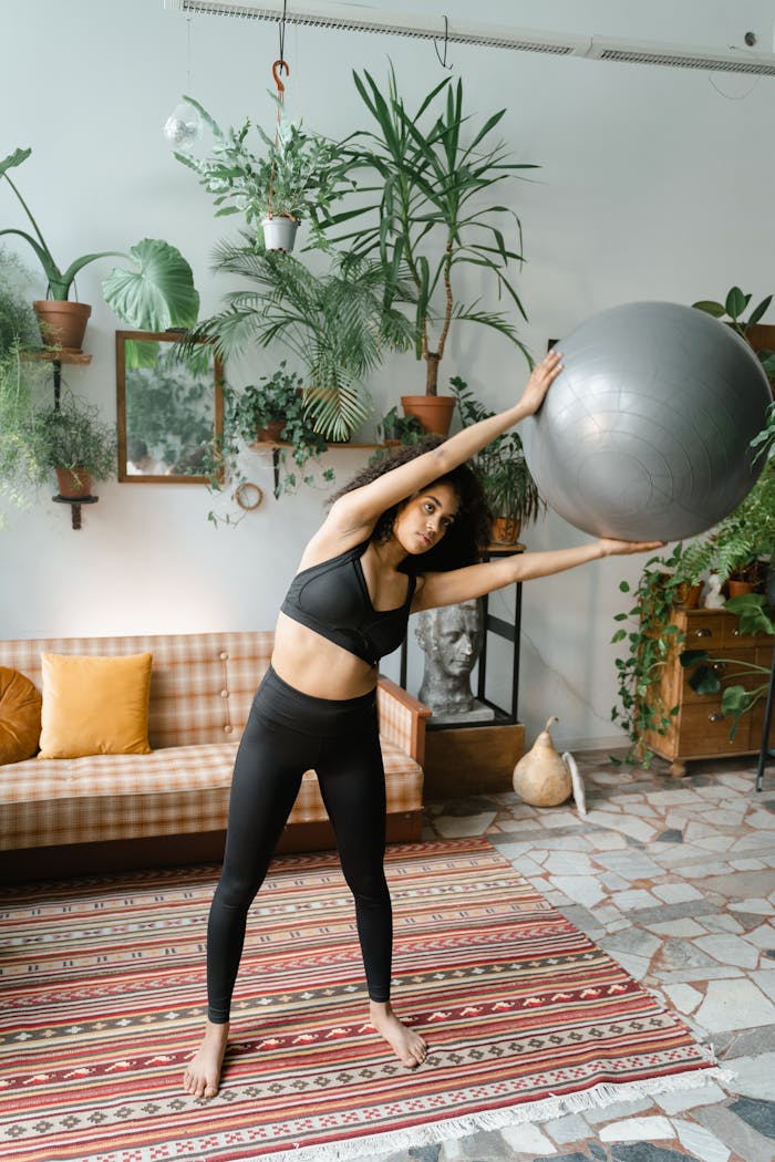 A woman in activewear performs fitness exercises with a yoga ball indoors, surrounded by greenery.